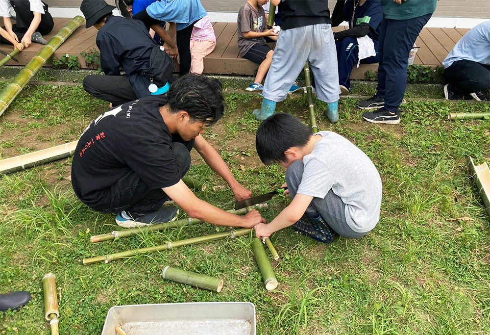 【6月農学校　流しそうめん】学生が竹の棒を押さえ、子どもがノコギリでその竹を切る様子。草の上でみんなで作業している。