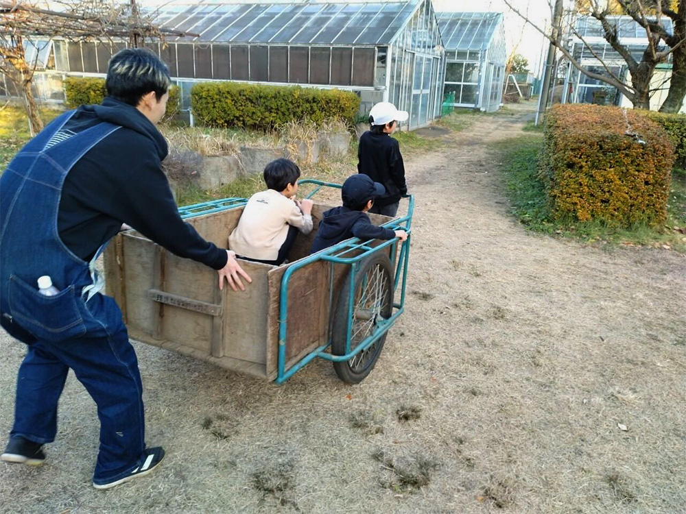 リアカーの後ろを押す学生と、リアカーを引っ張るこども、リアカーに乗っている子どもの後ろ姿