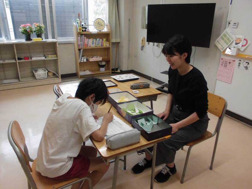 教室で先生が１人の児童に日本語を教える様子。先生は机を挟んで椅子に座り子どもと向き合って教えている。