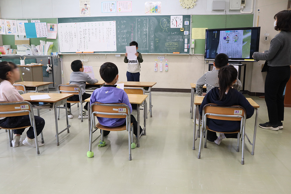 国際教室１年生の学期末の授業の一コマ。１人の児童が前に立って発表している様子。先生と、席に座る５人の児童が発表を聞いている。